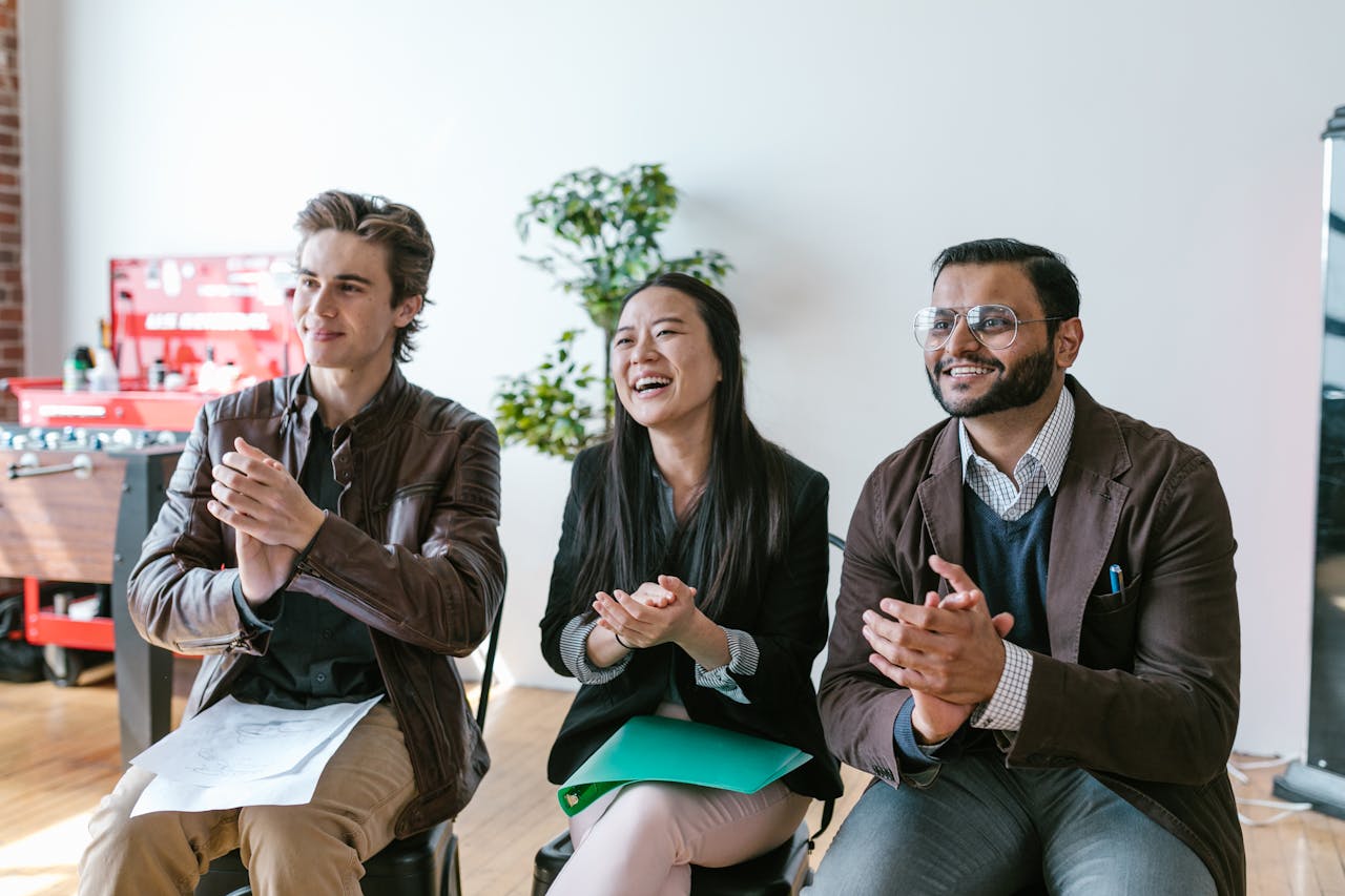 why-choose-us Three diverse team members clapping and smiling during an indoor meeting, showing positivity and teamwork.